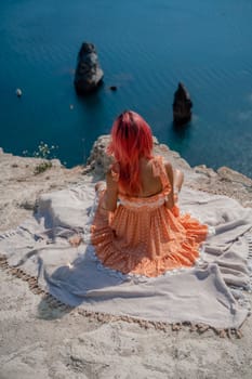 A girl with red hair sits with her back to the viewer on a picnic blanket in an orange dress. Summit on the mountain against the background of the sea and rocks in the sea.