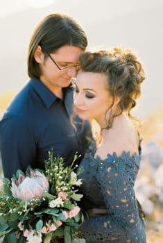 Groom hugs bride in a blue lace dress with a bouquet of flowers