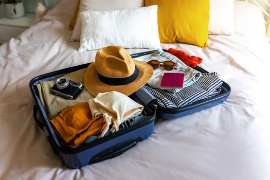 Open suitcase on the bed full of clothes and summer items: hat, sunglasses, swimsuit, passport and camera.