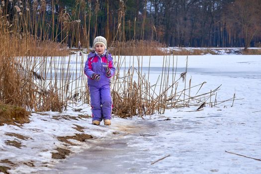 A cute child in a lilac jumpsuit walks on the lake in a winter park
