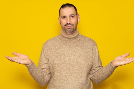 So what, who cares. Unsure and doubtful hispanic man with a beard wearing a beige turtleneck sweater isolated over yellow background. Indecision concept