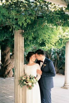 Groom hugs bride from behind by the shoulders near the column in the garden