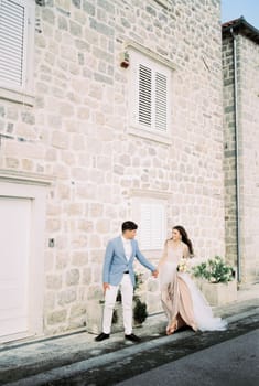 Bride and groom walk hand in hand past an old stone building