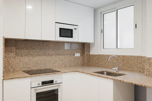 Modern kitchen is well lit by daylight from a sliding sash window. Beige granite walls with black spots. Kitchen furniture made of wood covered with white glossy paint. Built-in modern technology.