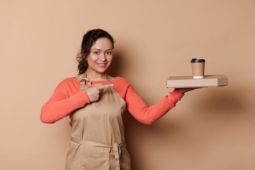 Pleasant multi-ethnic waitress points at takeaway food and drink in disposable containers, isolated on beige background