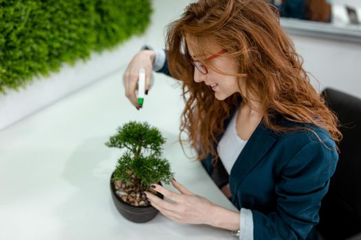 Top view of a young business woman who is working in the office with wall covered of grass. She sprays water on bonsai tree and smiles.