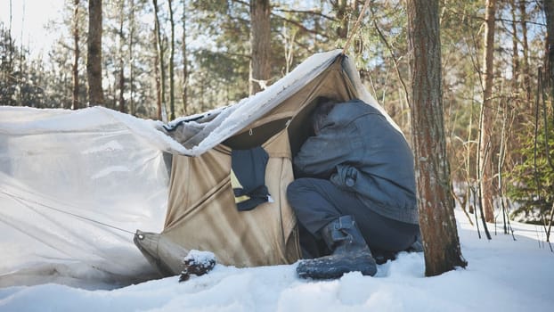 A homeless man climbs in and out of a tent in the woods in winter.