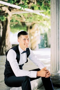 Smiling groom in a black vest sits on the steps near the column in the garden