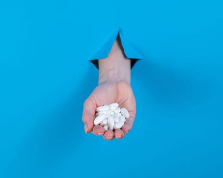 A woman is holding a handful of pills. A woman's hand sticks out of a hole in a cardboard blue background.