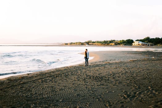 Girl in a sweater walks along the beach and looks at the surf