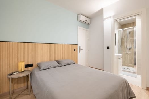 Bedroom with spacious bed against wall demarcated with wood paneling and blue paint and bedside table. Bedding is in beautiful gray color. From room there is doorway to bathroom with glass shower.