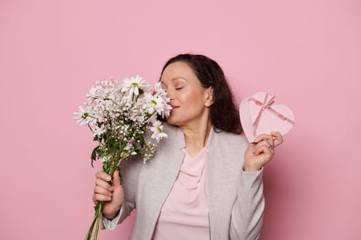 Delightful middle-aged woman sniffing beautiful bouquet of white chrysanthemum for Womens day, isolated pink background