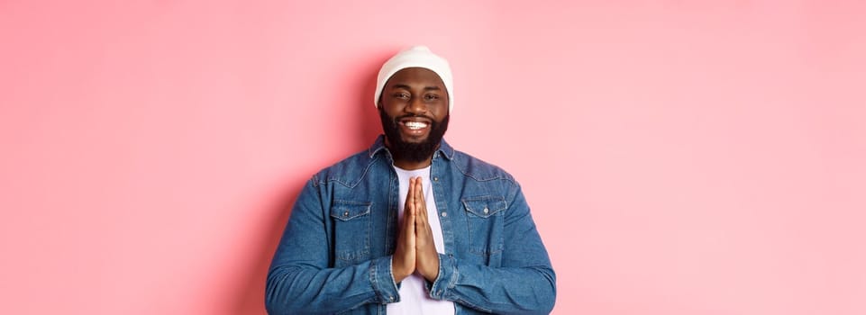 Happy smiling Black man saying thank you, holding hands in pray or namaste gesture, standing grateful against pink background