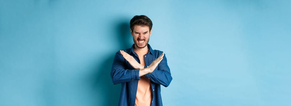 Angry young man frowning and clenching teeth outraged, showing cross gesture to stop or forbid something, standing on blue background