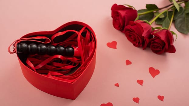 Valentine's Day gift for adults. Bdsm whip in a box in the shape of a heart and a bouquet of scarlet roses on a pink background