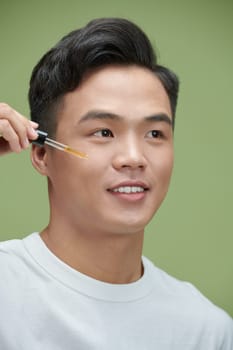 close up of young asian man applying moisturizing serum isolated on green