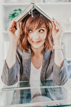 Woman book on her head. Surprised happy brunette in a jacket with a book on her head looks away against the backdrop of a bright office.