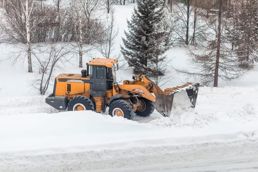 23.01.2023, Kemerovo, Russia. Orange tractor cleans up snow from the road and loads it into the truck. Cleaning and cleaning of roads in the city from snow in winter. Snow removal after snowfall