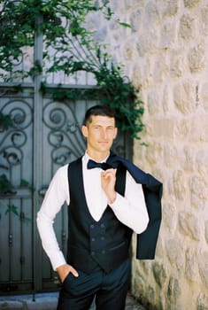 Groom in a vest with a jacket on his shoulder stands in the courtyard of an old stone building
