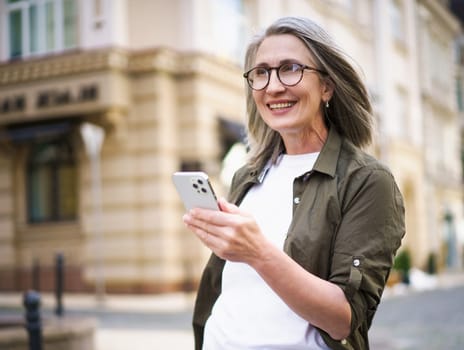 Joyful senior woman with grey hair and mobile phone in hand, enjoying her time in beautiful European city. Image showcases active and modern urban lifestyle of the elderly generation.