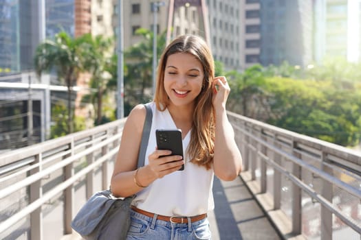 Attractive smiling woman walking on footbridge checking phone in the modern sustainable metropolis of Sao Paulo, Brazil