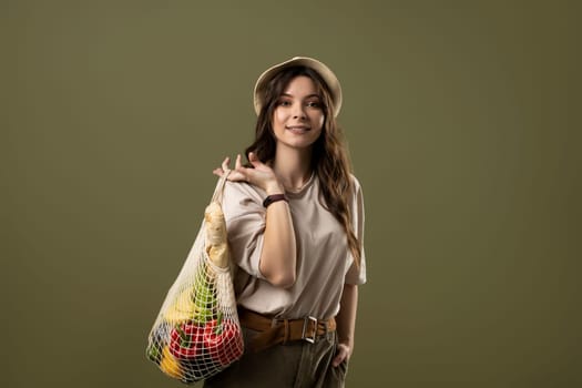 Portrait of brunette woman holding cotton net shopper, reusable mesh shopping bags with vegetables, products on pastel green background. Eco friendly mesh shopper. Zero waste, plastic free concept.