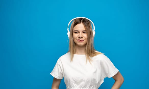 Beautiful attractive young blond woman wearing white t-shirt and glasses in white headphones listening music and smiling on blue background in studio. Relaxing and enjoying. Lifestyle.