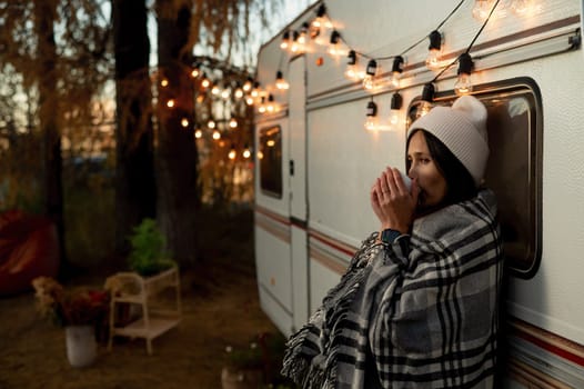 Caucasian woman in a knitted hat wrapped in a plaid and drinks warming tea outdoors. Travel in a camper in the fall.