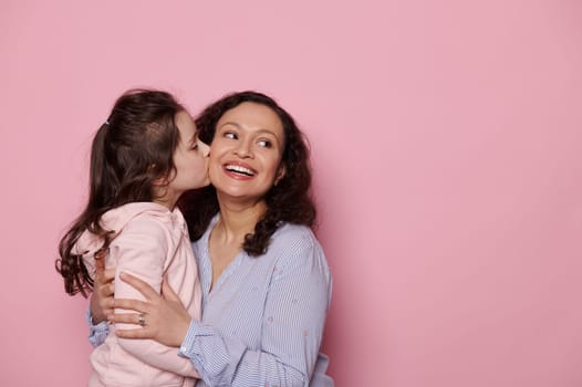 Caucasian cute little girl kissing her mother who smiles and looks aside at copy ad space on isolated pink background