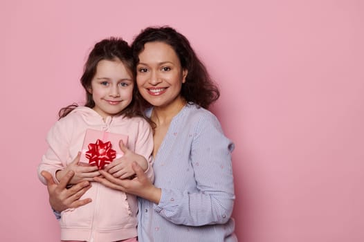 Loving mother and daughter embracing, holding a cute gift box and smiling looking at camera on isolated pink background