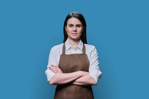 Portrait of confident young woman worker in apron on blue background