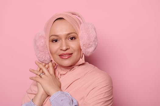 Beautiful Muslim woman, wearing a pink hijab and fluffy earmuffs, smiling looking at camera, isolated on pink background