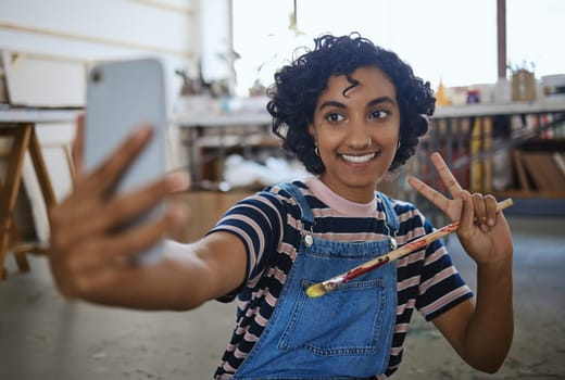 Indian woman artist, selfie with paint brush and art on floor of studio workshop to share to social media. Portrait of happy, creative professional painter and smiling after oil or watercolor project