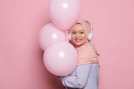 Lovely Muslim woman in pink hijab, a happy birthday girl with helium balloons, smiles at camera, enjoys festive occasion