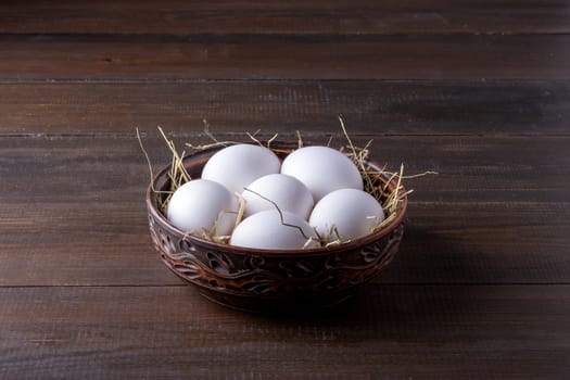 White raw chicken eggs lie on hay in a burnt clay bowl