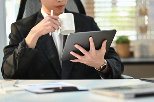 Cropped image of male business executive drinking coffee and reading business email on digital tablet in the morning.