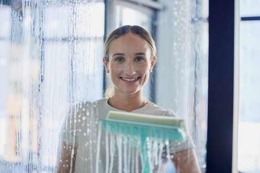 Cleaning, window and glass with a woman using a tool in her home with water, soap or foam inside. Housework, smile and service with a young female cleaner wiping a transparent pane in a modern house