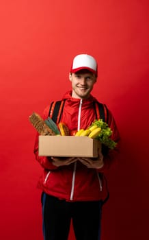 Fast young delivery man in red uniform carrying package box of grocery food and drink from store, market on red studio background. Delivery Concept.