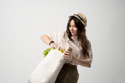 Smiling young woman in beige t-shirt and a hst with a mesh eco bag full of vegetables and on a white studio background. Sustainable lifestyle. Eco friendly concept. Zero waste.