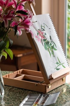 The easel with still life with purple lilies watercolor on the table prepared for painting