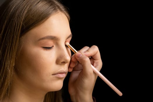 Make-up artist applying eyeshadow in the corner of model eyes and holding a shell with eyeshadow in a black studio. Professional make up.