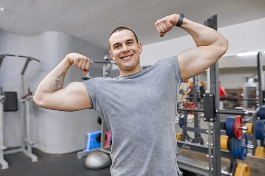 Young strong smiling muscular man in gym showing strong muscular arms.