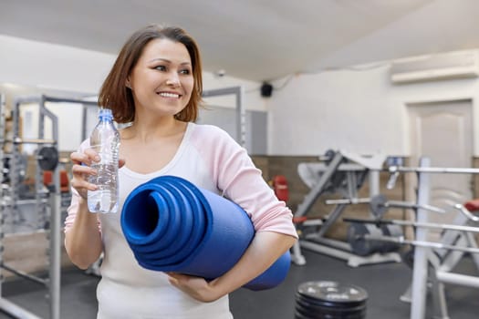 Portrait of smiling mature woman with bottle of water and sports mat in health club.