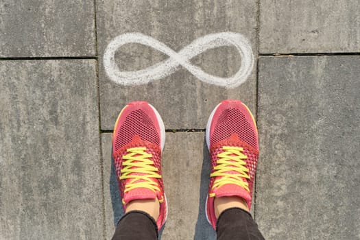 Infinity sign on gray sidewalk with woman legs in sneakers, top view