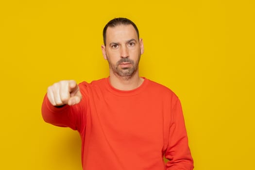 Bearded Hispanic man in a red sweater pointing at the camera with his finger, he is very serious and has an accusatory attitude. Isolated on yellow studio background