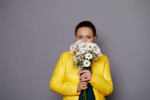 Portrait of a charming dark-haired woman with a bouquet of flowers, looking at camera, isolated over gray background.