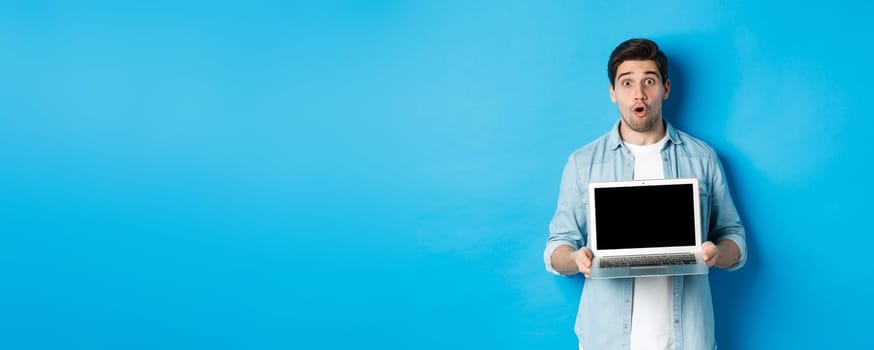 Man showing advertisement on laptop screen and looking amazed, saying wow and looking at camera, standing against blue background