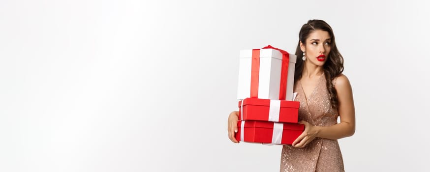 New Year, Christmas and celebration concept. Attractive young woman in elegant dress, looking away, carry gifts, standing over white background