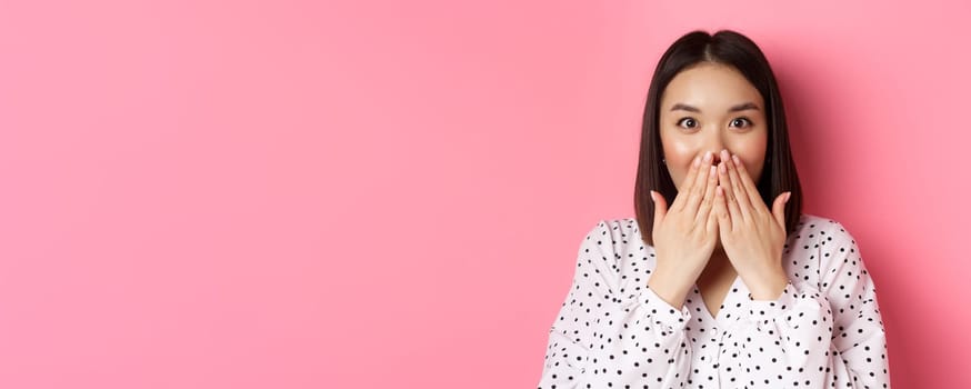 Close-up of beautiful asian girl looking surprised and excited, cover mouth and staring amazed at camera, standing over pink background