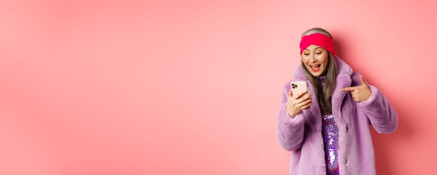 Online shopping and fashion concept. Cool asian senior woman checking out promotion on mobile phone, pointing finger at smartphone screen and smiling pleased, pink background.